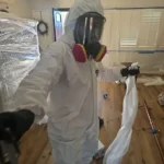 Technician in hooded coveralls and a full-face respirator with P100 cartridges inside a sealed containment, contents wrapped in plastic