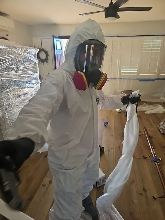 Technician in hooded coveralls and a full-face respirator with P100 cartridges inside a sealed containment, contents wrapped in plastic