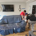 Two technicians wrapping a sofa in plastic where it stands before drying equipment is set