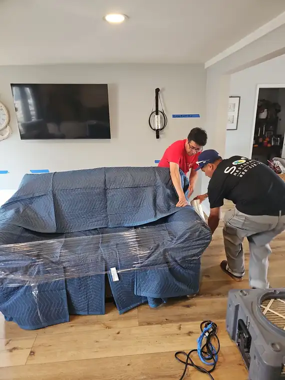 Two technicians wrapping a sofa in plastic where it stands before drying equipment is set