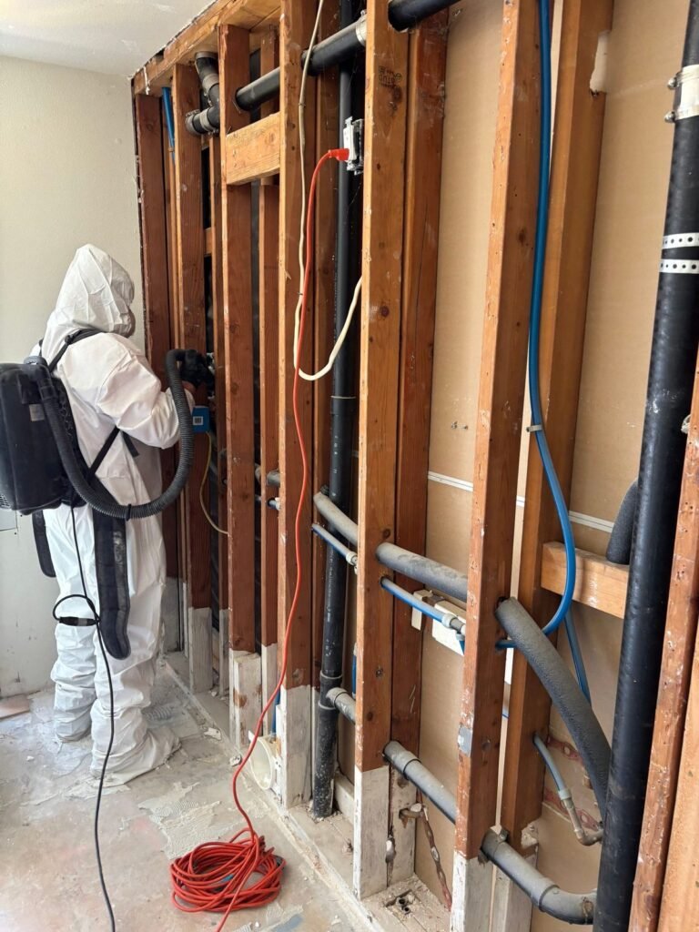 A technician with a backpack vacuum at an open wall with pipework exposed