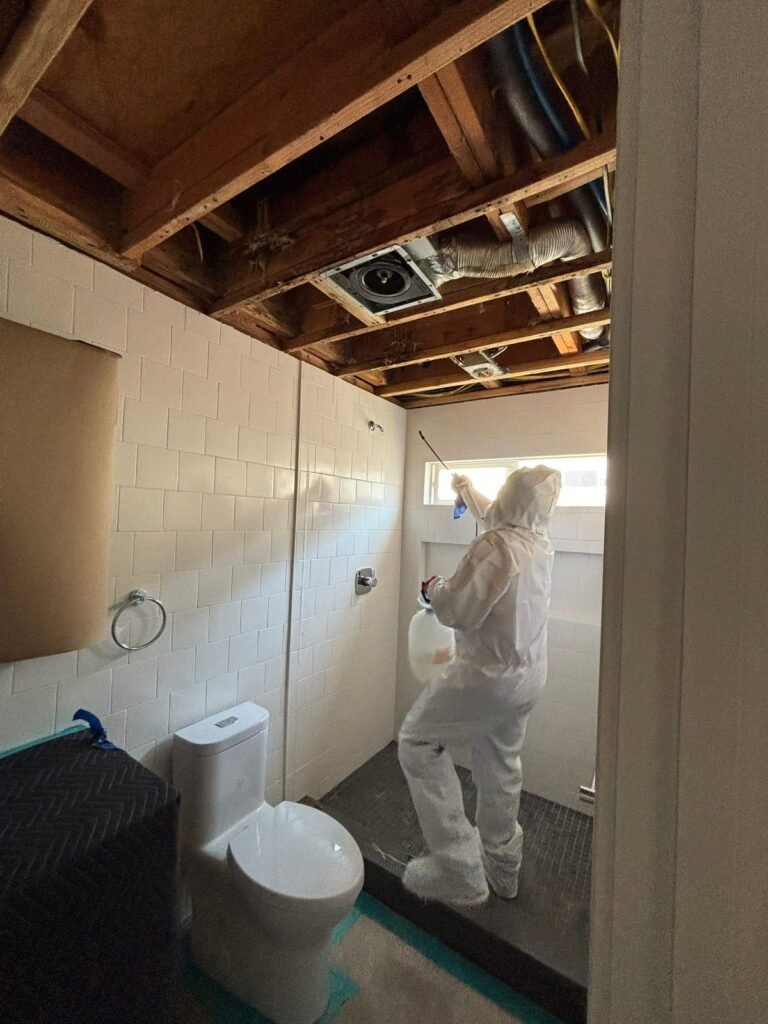 A bathroom with the ceiling opened to the joists and a technician working the wall