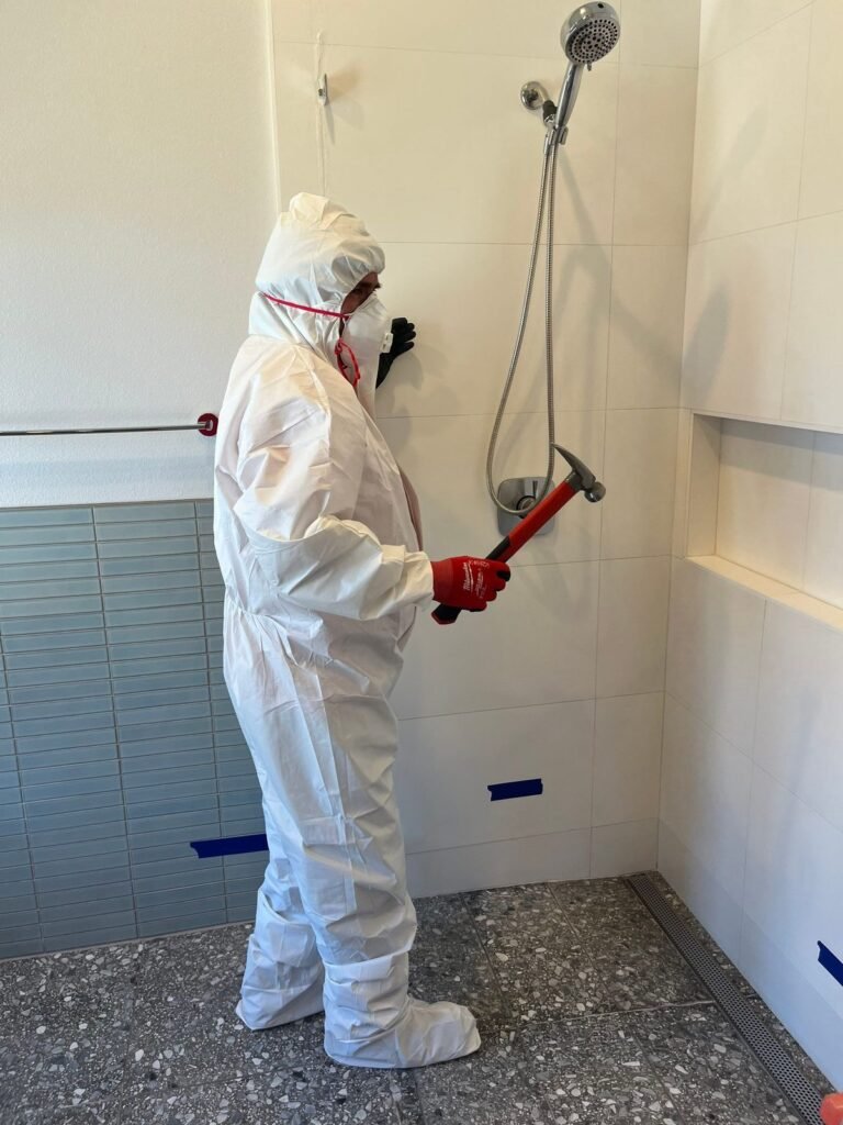 A technician using a pry bar on the wall beside a tiled shower