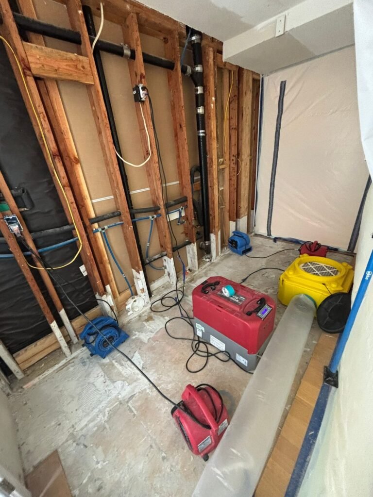 An opened wall with the pipes visible, a red dehumidifier and a yellow air mover working